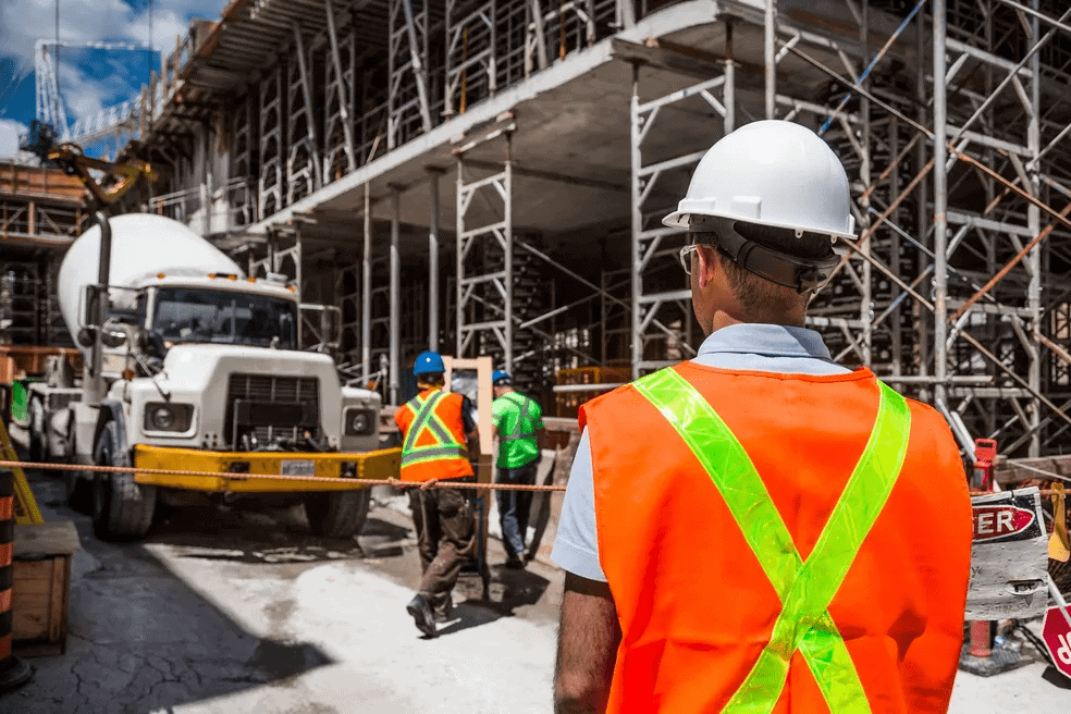 Homem em obra, com colete alaranjado e olhando para caminhão e trabalhadores em atividade. Imagem ilustrativa texto sobre franquia de construção