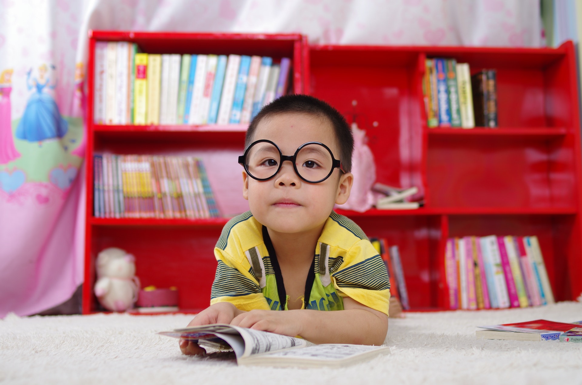 Criança usando um óculos deitada em um carpete em uma sala de aula com estantes vermelhas ao fundo. Imagem ilustrativa do texto franquia educacional infantil.