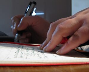 Duas mãos sobre um caderno com capa vermelha e uma delas segurando uma caneta e escrevendo. Imagem ilustrativa do texto plano de negócios para franquia.