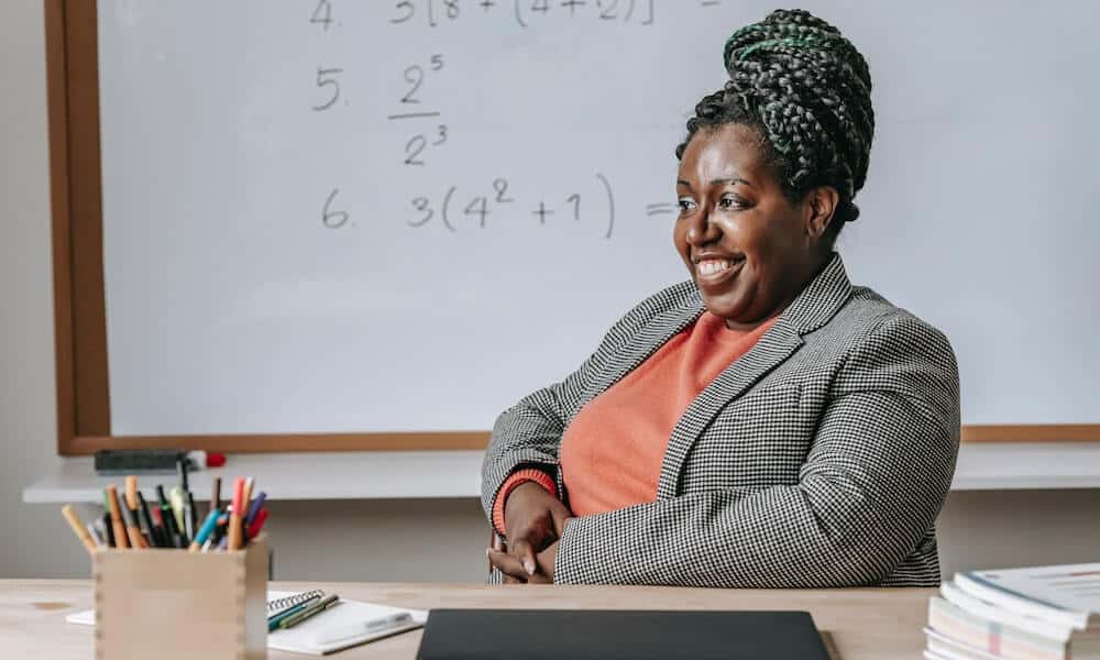 Mulher com cabelo trançado e casaco cinza sorri olhando para o lado. AO fundo, um quadro branco com fórmulas matemáticas. Imagem ilustrativa texto curso de matematica para criancas.