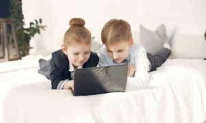 Duas crianças vestidas com roupa social, a menina de terninho preto e o menino de camisa social azul, olham para um notebook e sorriem. Imagem ilustrativa texto mentalidade empreendedora das criancas.