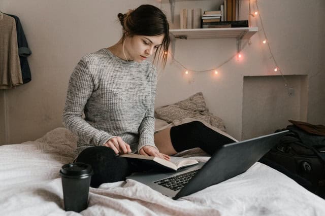 Mulher em cima da cama com livro aberto e notebook. Ilustração do texto sobre como montar um negócio.