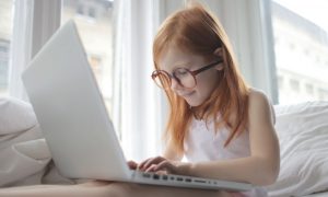 Menina de camisa branca digita em um laptop enquanto morde o lábio sorrindo. Imagem ilustrativa texto curso de desenvolvedor de games para crianças.