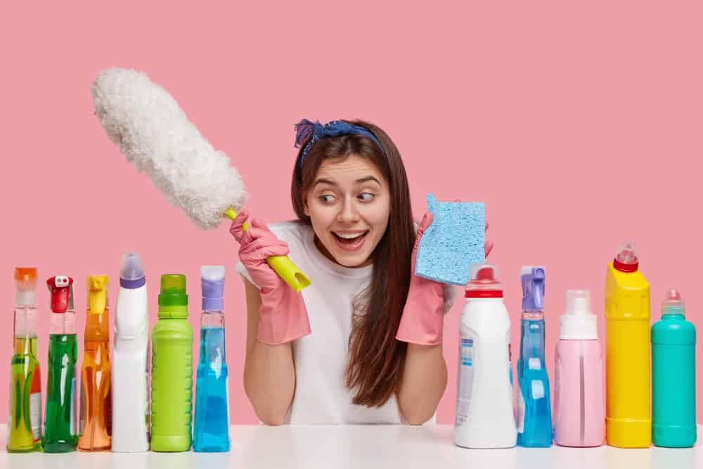 mulher com espanador e bucha nas mãos cercada por um fundo rosa e muitos produtos de limpeza. Imagem ilustrativa do texto sobre franquia de produtos de limpeza e higiene