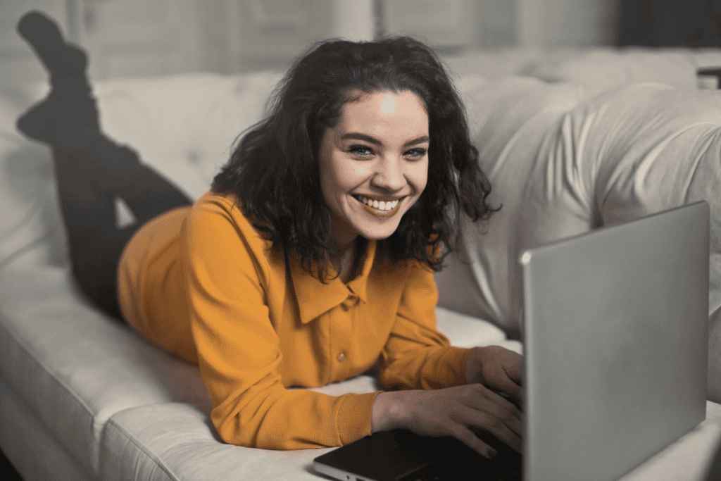 Mulher deitada de bruços no sofá e usando um notebok, sorrindo para a frente; ela usa uma blusa de frio amarela. Imagem ilustrativa texto sobre franquias mais rentáveis