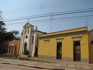 Imagem da frente de um comércio de cidade do interior com a parede amarela e duas portas ao lado da entrada de uma igreja com as paredes brancas. Imagem ilustrativa texto como abrir um negócio em cidade pequena.