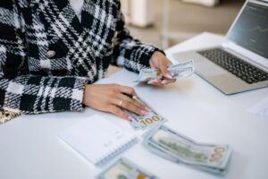 Pessoa em uma mesa contando notas de dinheiro ao lado de um computador. Imagem ilustrativa do texto como começar um investimento com pouco dinheiro.