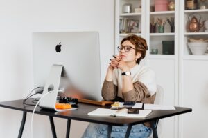 Mulher sentada em uma mesa de trabalho em casa, com um computador na sua frente e com as mãos cruzadas sobre o queixo. Imagem ilustrativa do texto sobre franquia para trabalhar meio período.