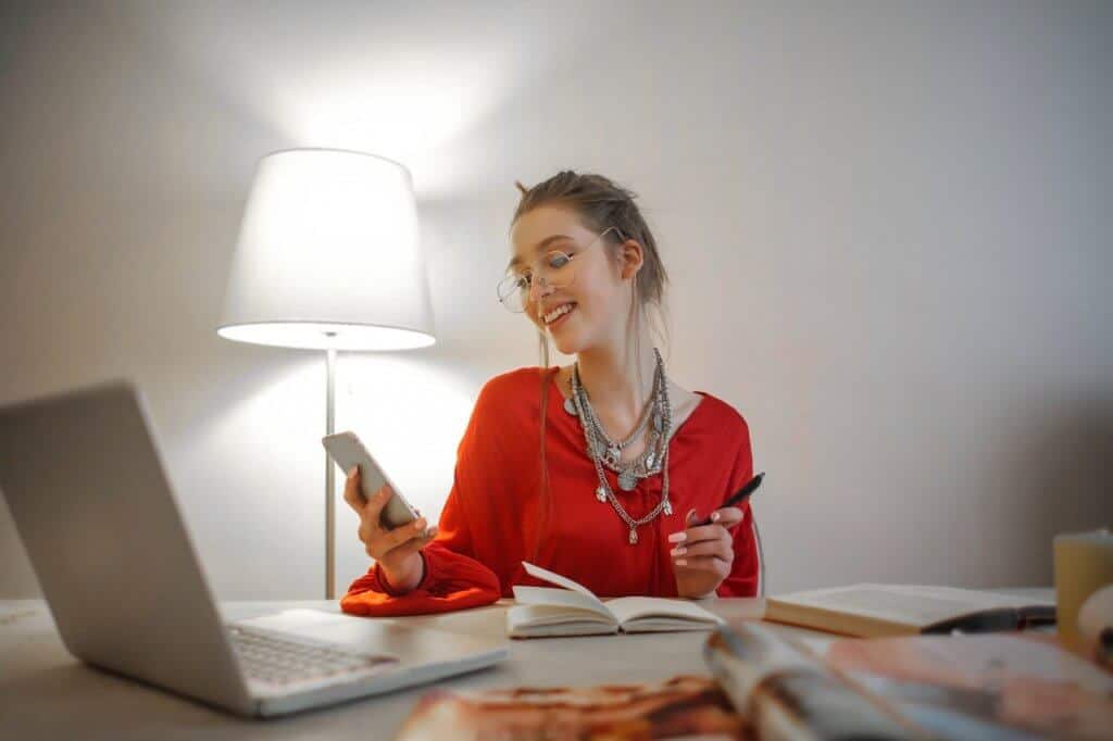 Mulher usando blusa vermelha e óculos sorrindo, segurando celular em uma mão e caneta em outra. Ilustração do texto sobre microfranquia home office.