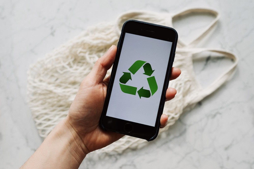 Mão segurando um celular preto com o símbolo de reciclagem em verde na tela. Ao fundo uma sacola branca. Imagem ilustrativa do texto negócios sustentáveis.