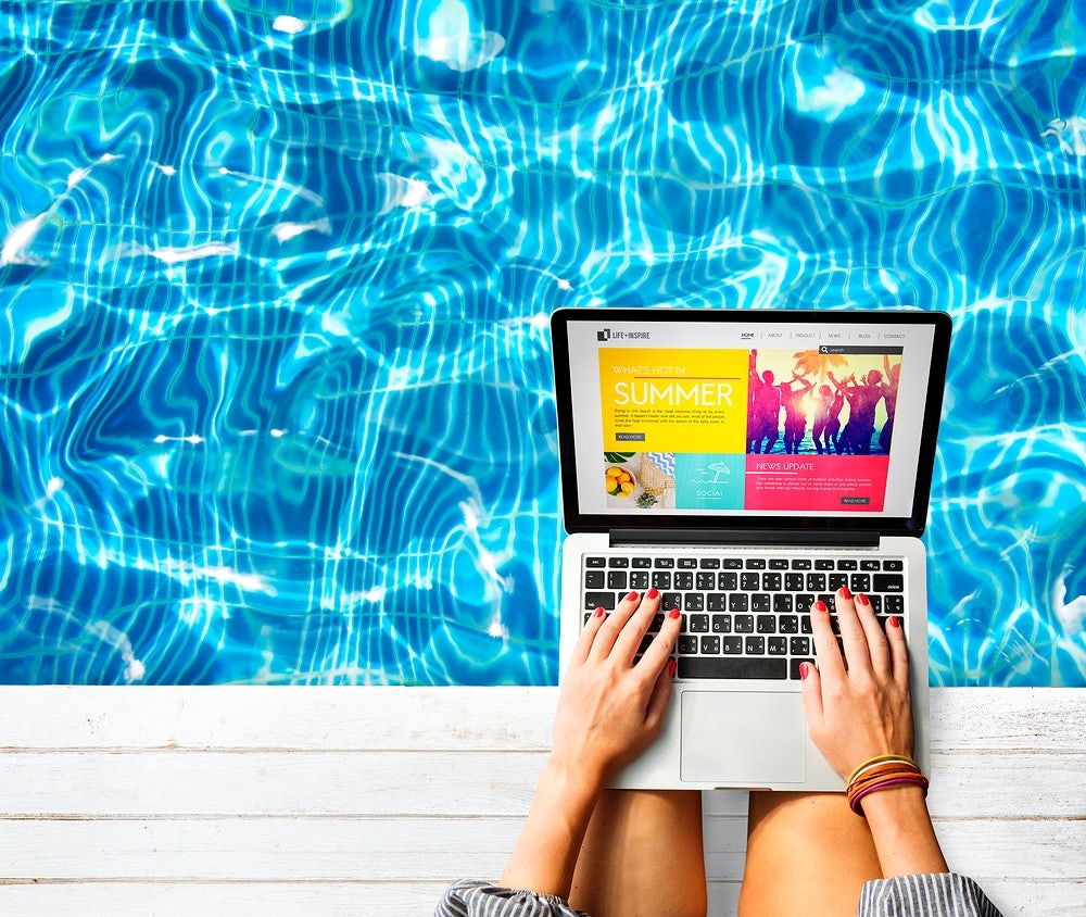 Pessoa mexendo em um notebook com imagens coloridas na tela e na beira de uma piscina azul. Imagem ilustrativa do texto agência de viagens física X agência virtual qual modelo escolher.