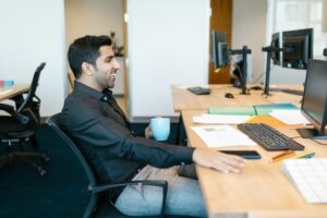 Homem sentando em uma cadeira de escritório usando uma camisa social escura olhando para a tela de um computador e sorrindo. Na mesa vemos diversos itens de escritório e um teclado. Imagem ilustrativa do texto investir em franquia.