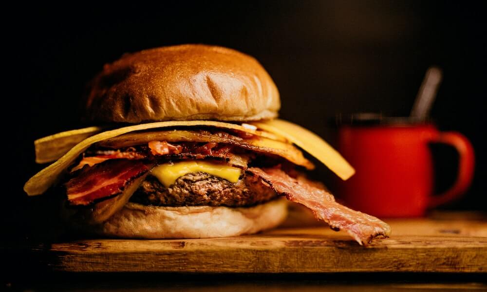 Imagem de hambúrguer com queijo e fatias de bacon caídas. Imagem ilustrativa franquia de hamburgueria.