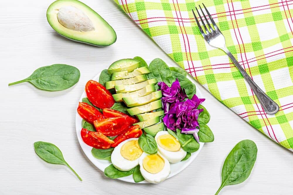 Prato de salada ao lado de abacate, talheres e forro de mesa xadrez em tons verde e laranja. Ilustração do texto sobre franquias de alimentação saudável.