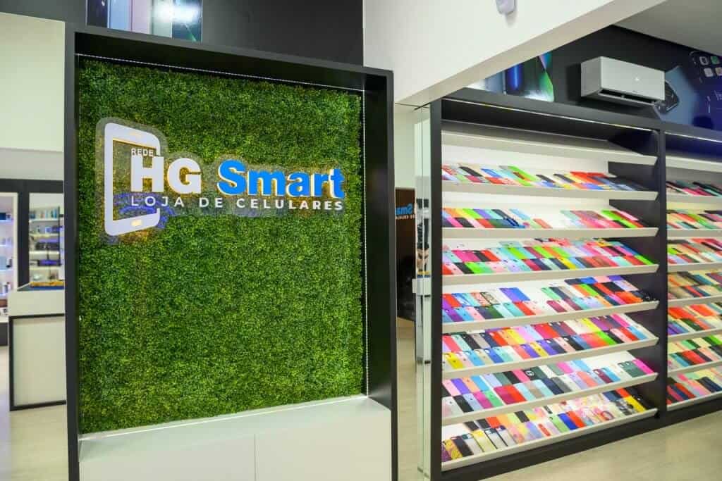 Logomarca da HG Smart na parede. A parede tem folhagem verde e a logomarca tem as cores branca e azul. Ao lado, há uma prateleira com capinhas coloridas para celular.