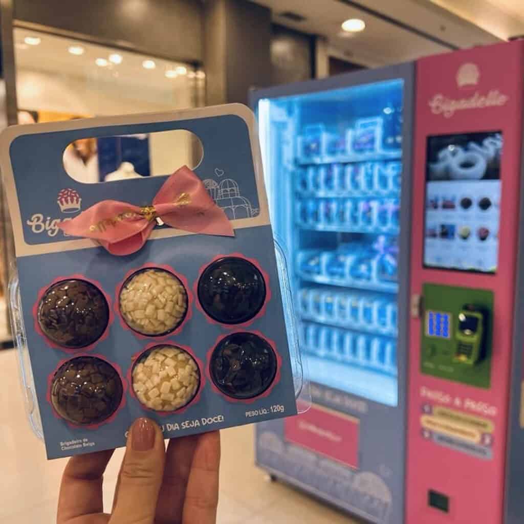 Uma mão mostra uma embalagem de brigadeiros em frente a uma vending machine da franquia Bigadello.